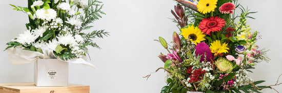 two bouquets for flowers and gifts for mothers day, one white and green in a white vase and the other yellow, red and white with greens set against a white wall