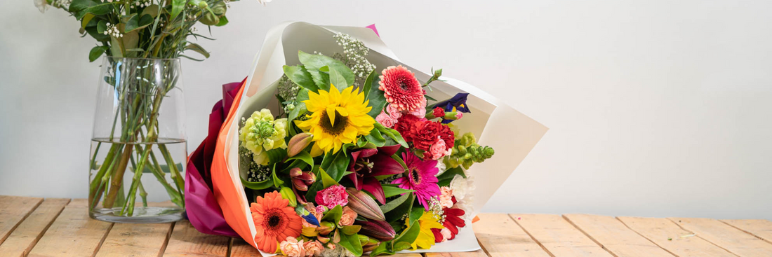 arrangement of brightly hued flowers wrapped in hot pink paper and sitting next to greenery in a glass vase both on a wooden slatted table