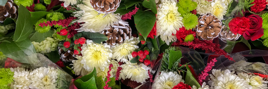 close up of lots of flowers including red roses, white lisianthus, reen an dpink blooms, greenery and red berries scattered with white tipped pine cones