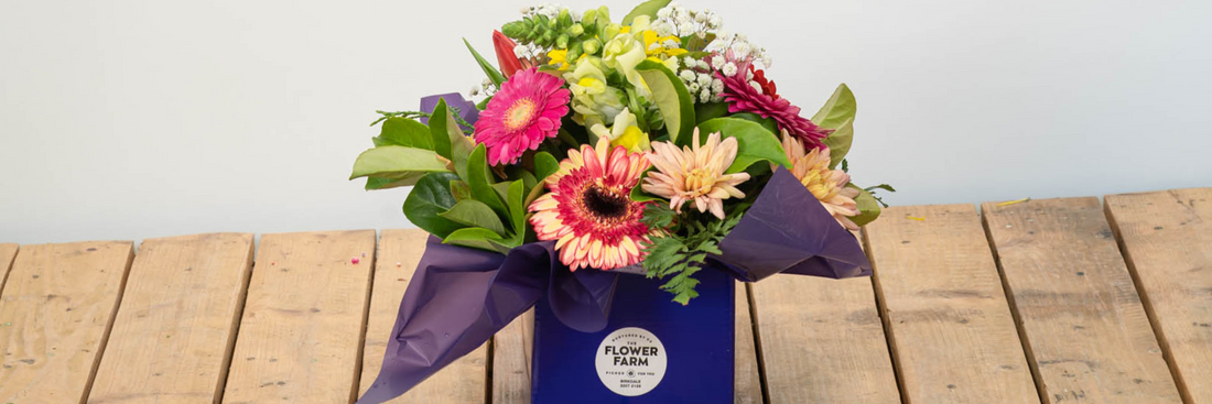 gorgeous and brightly coloured flower farm arrangement in a purple box with a logo sitting on a slatted wooden table