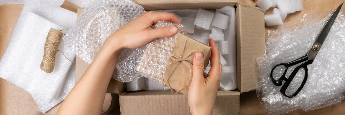 close up of a woman's hands bubble wrapping a brown paper package to put into a larger brown box with scissors and bubble wrap and tissue paper scattered around