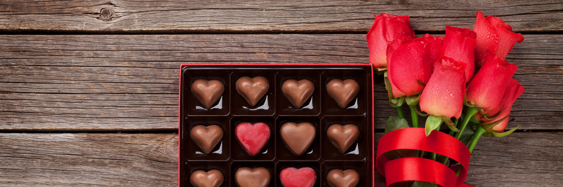 square box of heart shaped chocolates next to a bunch of red roses wrapped in red ribbon on a wooden table